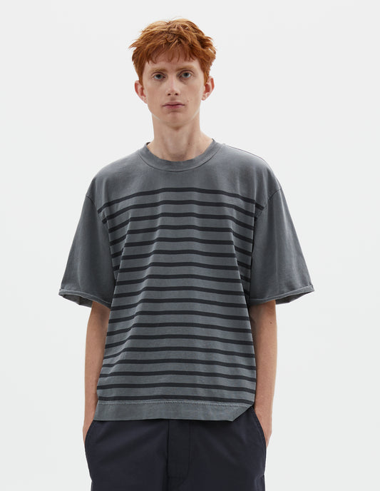 Cropped body image, front view image of model wearing blueish grey short sleeve t shirt with black stripes, flatlock stitch seam detailing and rolled raw sleeve hems held with a single bar tack. Styled with dark navy blue trousers.
