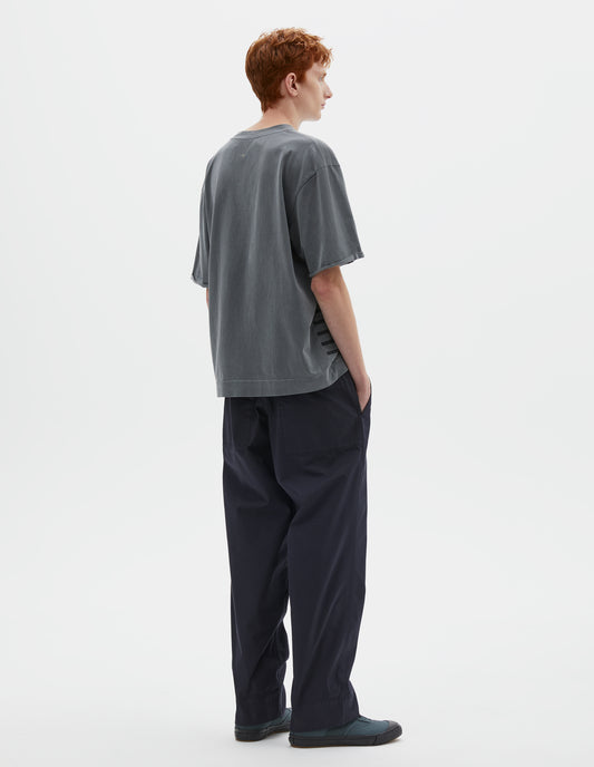 Full body image, back view of model wearing blueish grey short sleeve t shirt with flatlock stitch detailing and rolled raw sleeve hems with a single bar tack. Styled with dark navy blue trousers and blue high top canvas trainers. 
