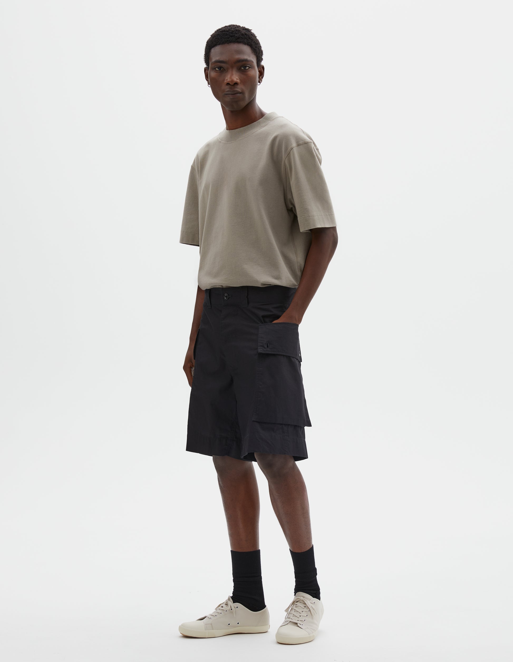 Full body image, front view of model wearing black cotton shorts with cargo pockets, belt loops and corozo brace button closure. Styled with beige t-shirt, black socks and off white canvas trainers. 

