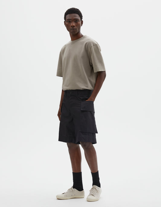 Full body image, front view of model wearing black cotton shorts with cargo pockets, belt loops and corozo brace button closure. Styled with beige t-shirt, black socks and off white canvas trainers. 
