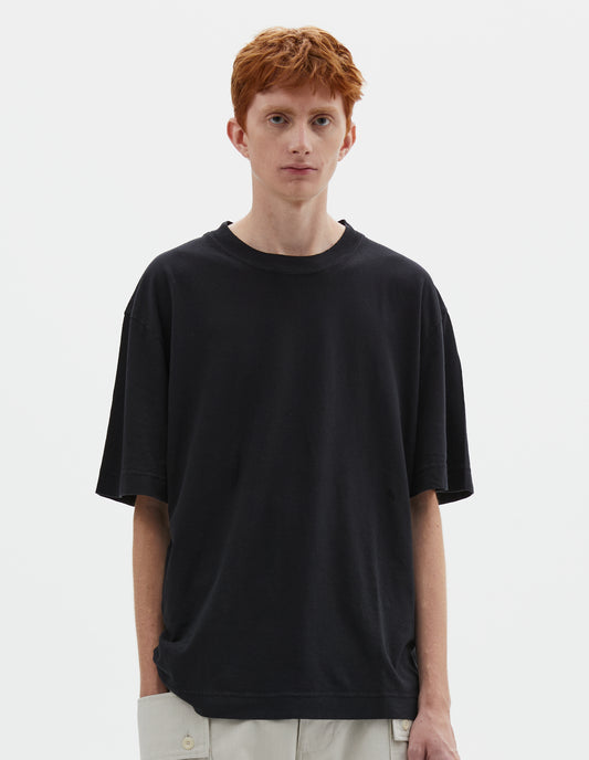 Cropped body image, front view of model wearing boxy black  t-shirt with herringbone tape detailing on inside neck and vent split hem. Styled with off white trousers.