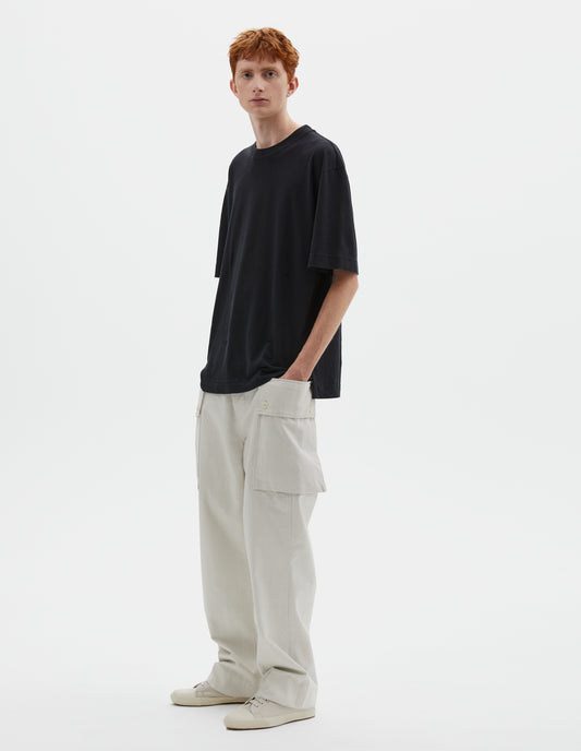 Full body image, front view of model wearing boxy black  t-shirt with herringbone tape detailing on inside neck and vent split hem. Styled with off white trousers and beige canvas trainers.