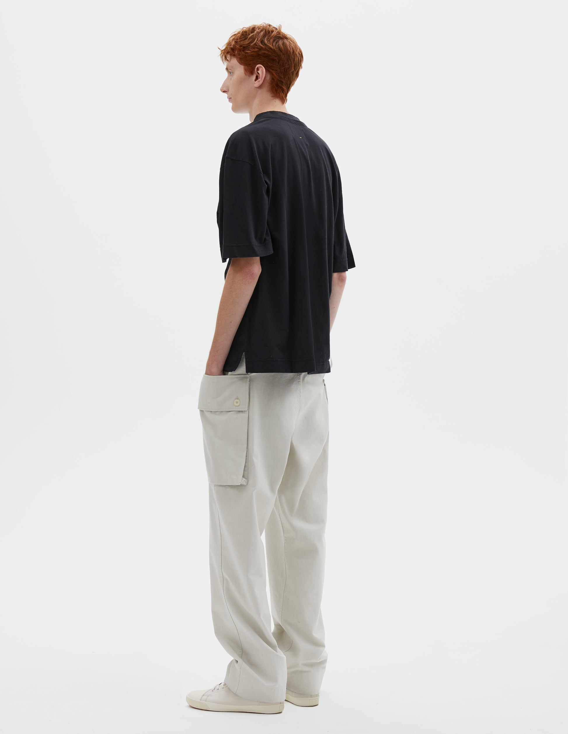 Full body image, back view of model wearing boxy black t-shirt with vent split hem. Styled with off white trousers and beige canvas trainers.