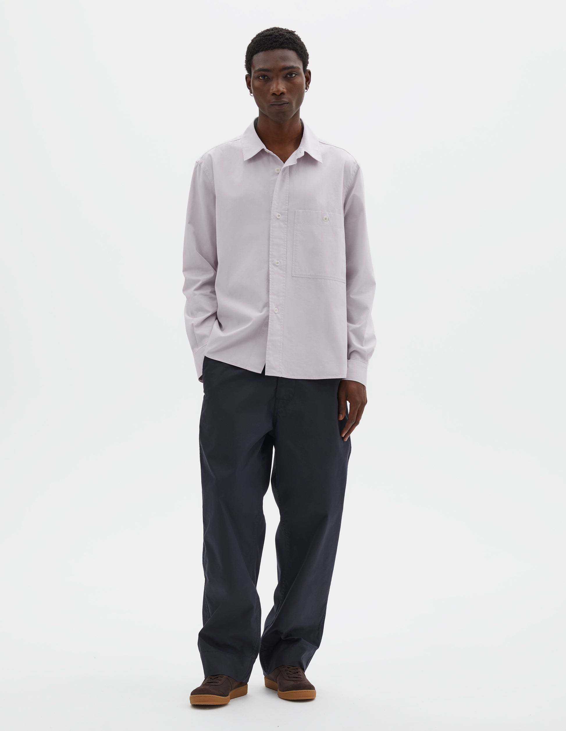 Full body image, front view of model wearing light grey cotton canvas shirt, with raised placket, corozo button fastening and patch pocket on chest. Styled with navy blue trousers and brown suede trainers.