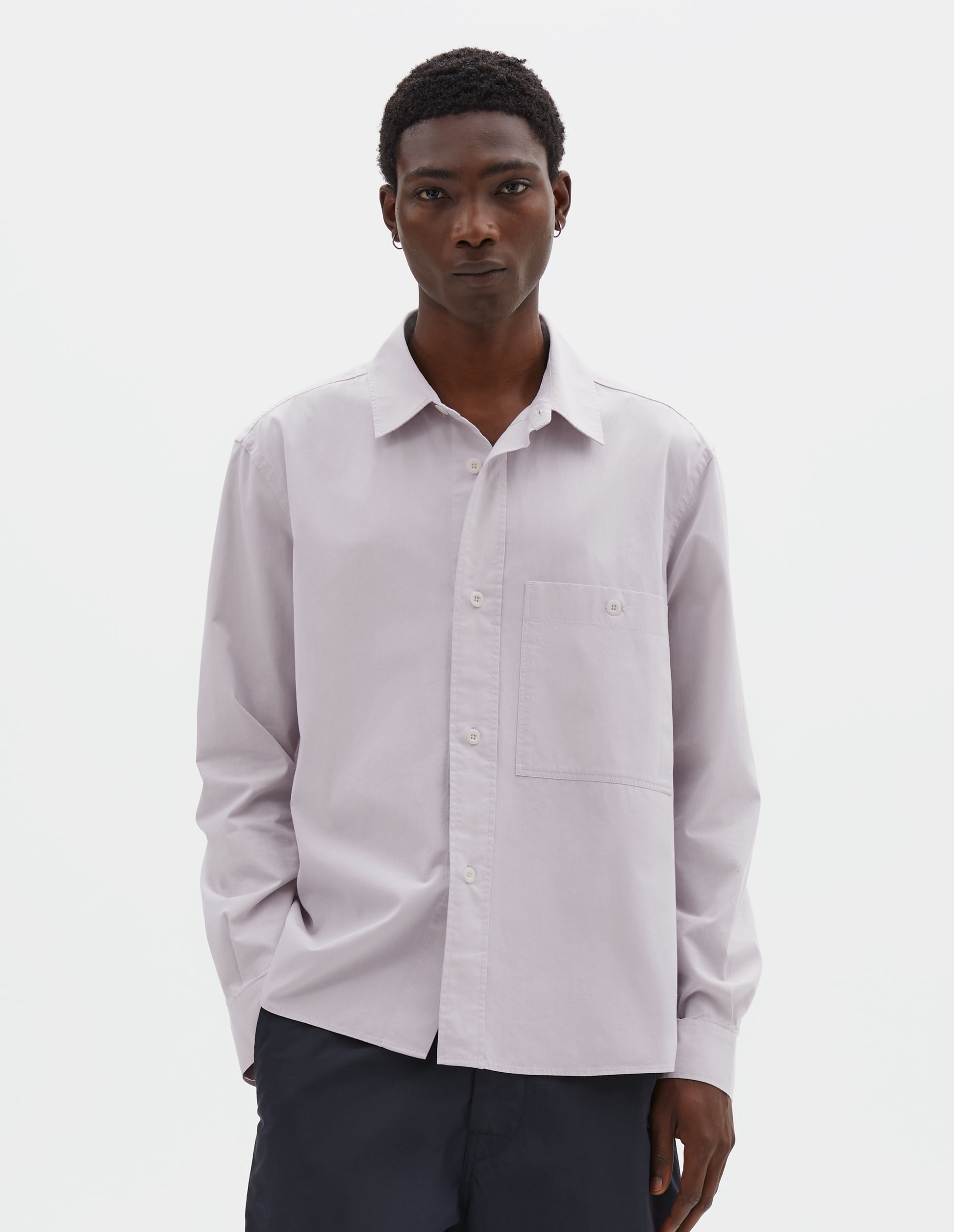 Cropped body image, front view of model wearing light grey cotton canvas shirt, with raised placket, corozo button fastening and patch pocket on chest. Styled with navy blue trousers.