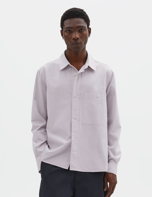 Cropped body image, front view of model wearing light grey cotton canvas shirt, with raised placket, corozo button fastening and patch pocket on chest. Styled with navy blue trousers.