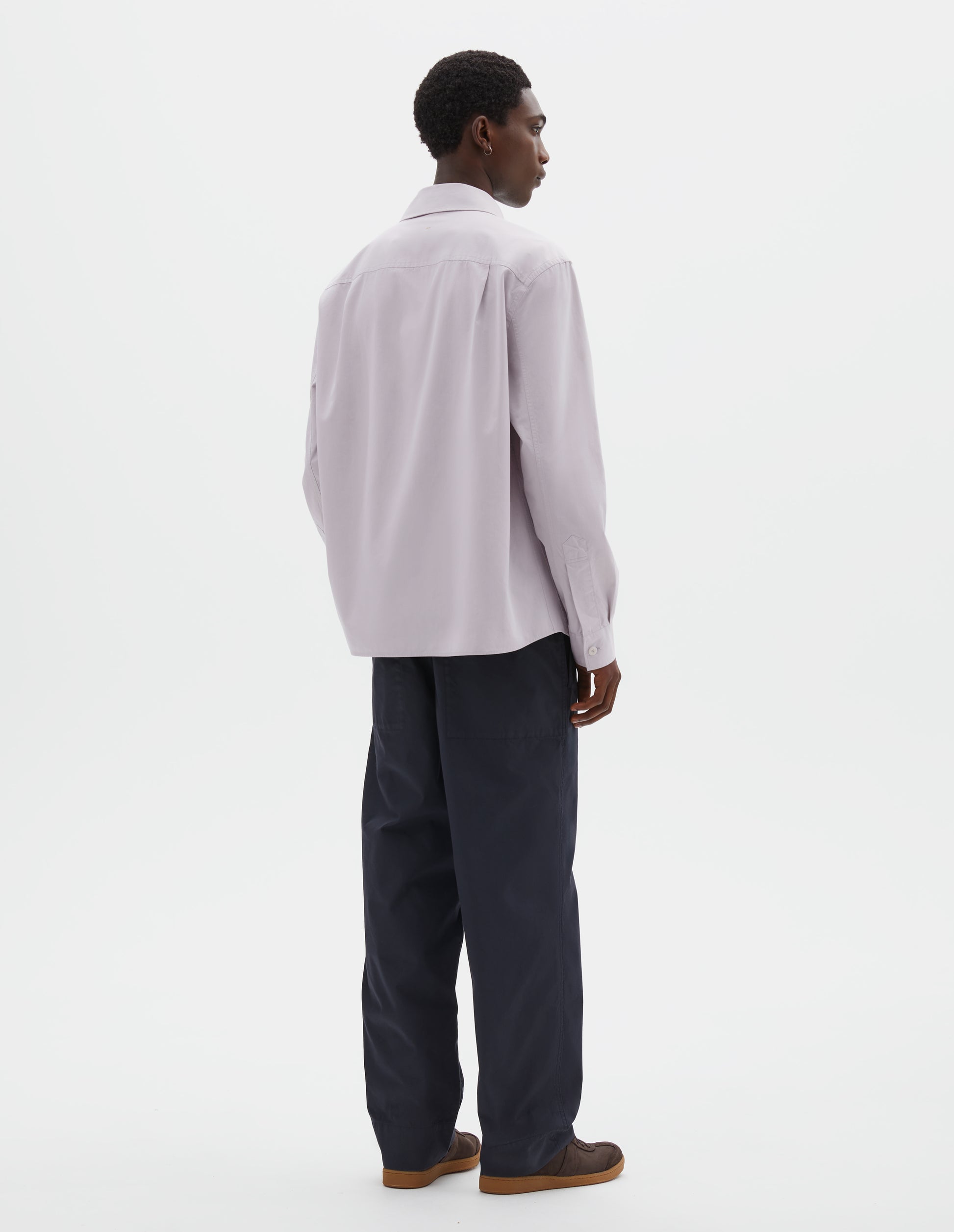 Full body image, back view of model wearing light grey cotton canvas shirt, with corozo button cuffs and side pleats on back yoke. Styled with navy blue trousers and brown suede trainers. 