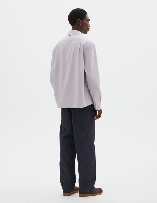 Full body image, back view of model wearing light grey cotton canvas shirt, with corozo button cuffs and side pleats on back yoke. Styled with navy blue trousers and brown suede trainers. 