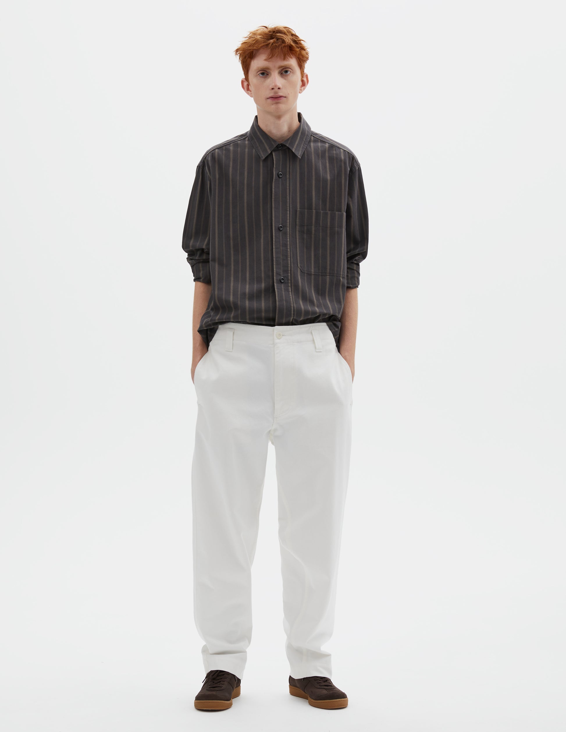Full body image, front view of model wearing white cotton drill trousers with belt loops, slant side pockets and corozo button fastening. Styled with grey, black and green striped shirt and brown suede trainers.