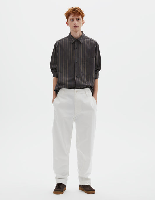 Full body image, front view of model wearing white cotton drill trousers with belt loops, slant side pockets and corozo button fastening. Styled with grey, black and green striped shirt and brown suede trainers.