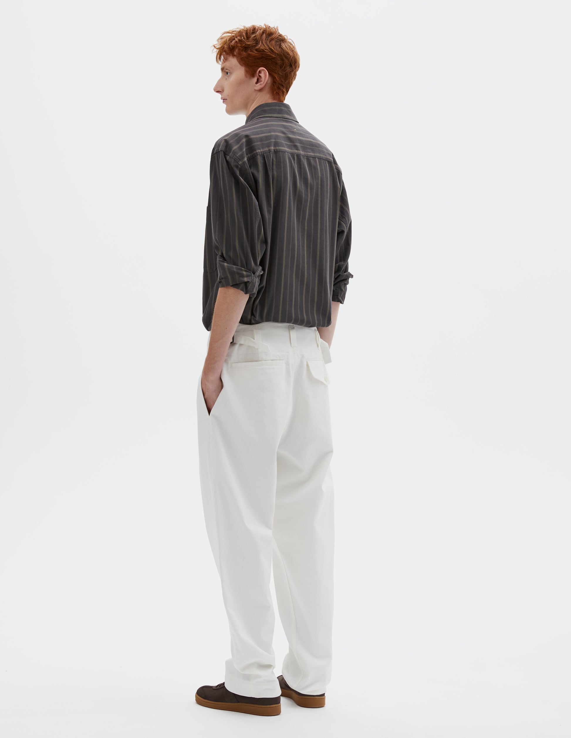 Full body image, back view of model wearing white cotton drill trousers with belt loops, side adjustors, welt back and flap back pockets. Styled with grey, black and green striped shirt and brown suede trainers.