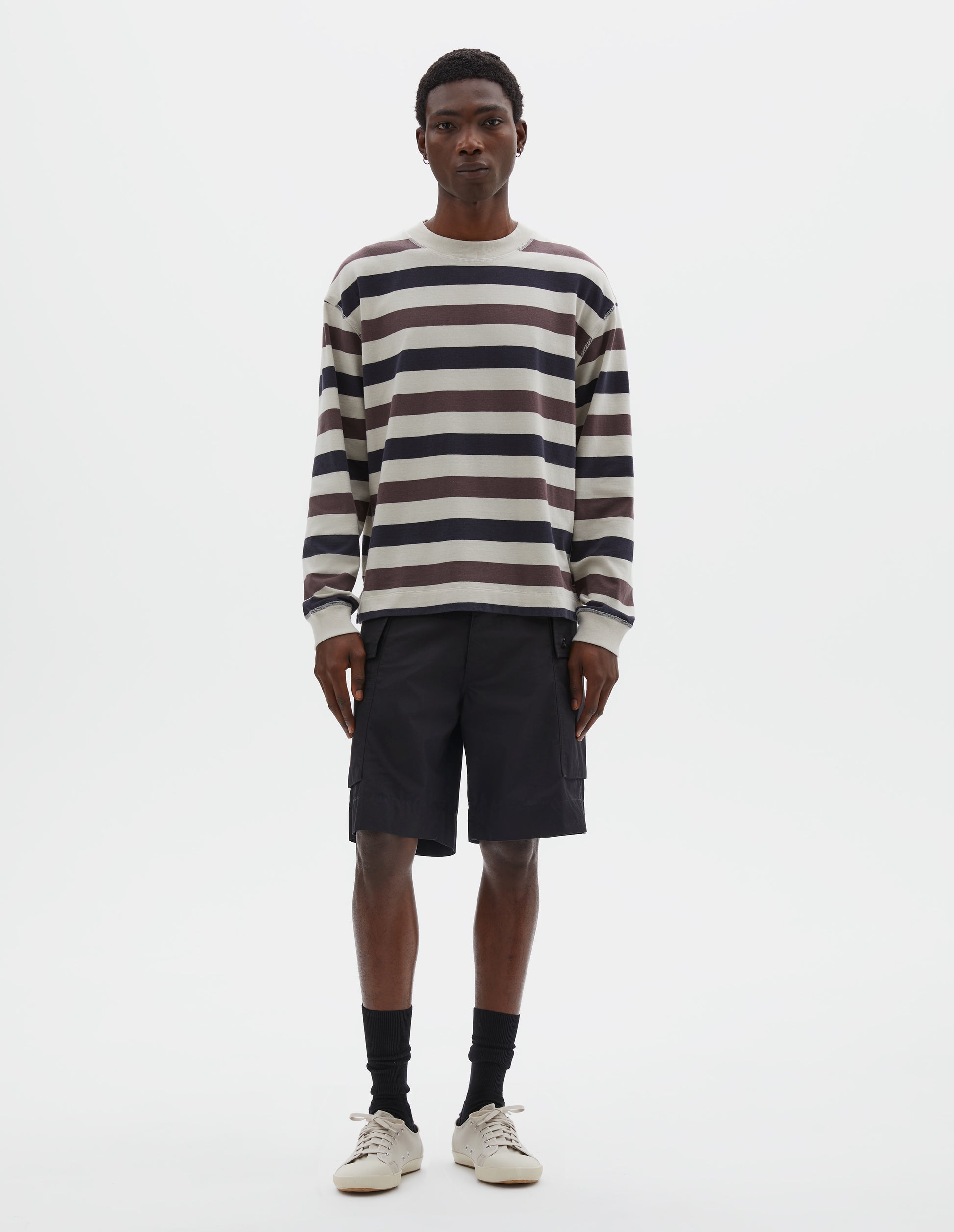 Full body image, front view of model wearing off white, dark navy and purple striped long sleeve top, with contrast rib on neckline and cuffs and flatlock stitch detailing. Styled with black shorts, black socks and beige canvas trainers.