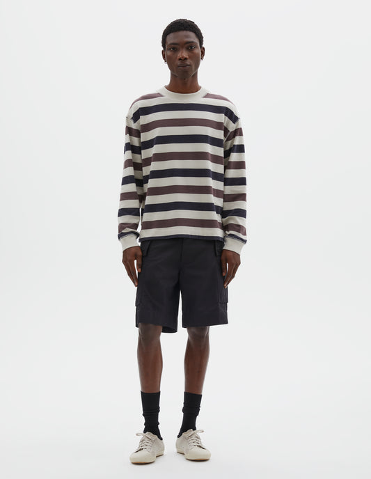 Full body image, front view of model wearing off white, dark navy and purple striped long sleeve top, with contrast rib on neckline and cuffs and flatlock stitch detailing. Styled with black shorts, black socks and beige canvas trainers.