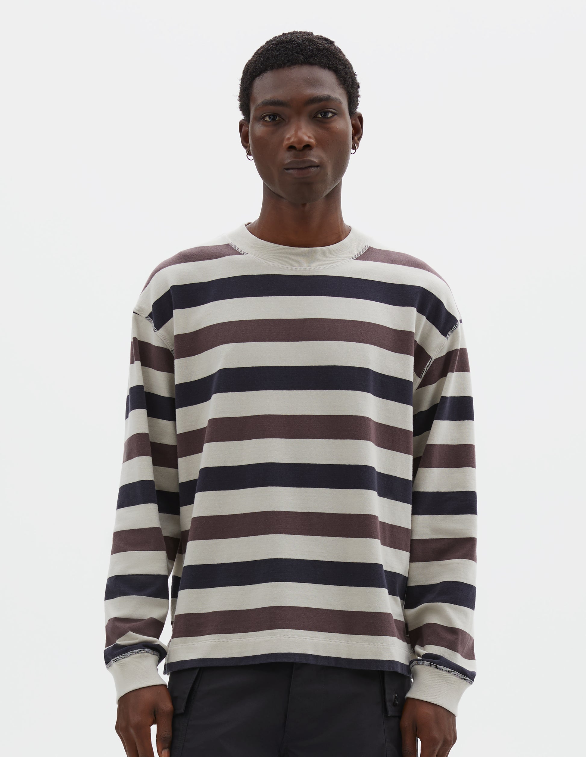 Cropped body image, front view of model wearing off white, dark navy and purple striped long sleeve top, with contrast rib on neckline and cuffs and flatlock stitch detailing. Styled with black shorts.