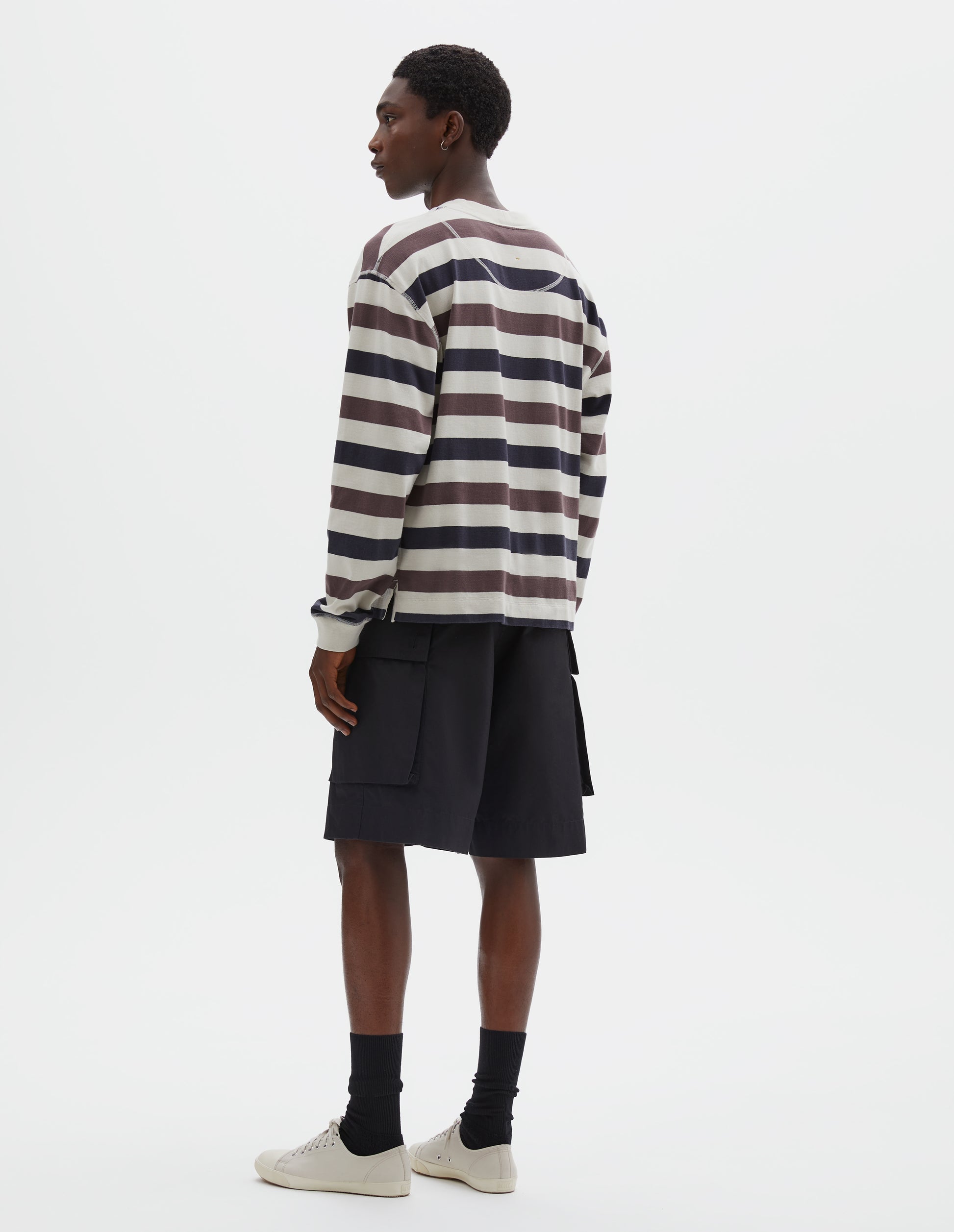 Full body image, back view of model wearing off white, dark navy and purple striped long sleeve top, with contrast rib on neckline and cuffs, flatlock stitch detailing and half moon neck lining. Style with black shorts, black socks and beige canvas trainers.