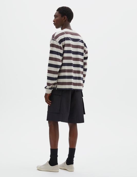 Full body image, back view of model wearing off white, dark navy and purple striped long sleeve top, with contrast rib on neckline and cuffs, flatlock stitch detailing and half moon neck lining. Style with black shorts, black socks and beige canvas trainers.