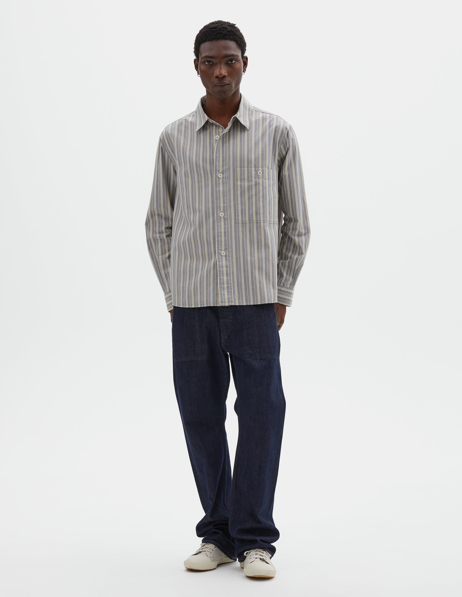 Full body image, front view of model wearing grey, white and yellow striped long sleeve cotton shirt, with dropped shoulder, raised placket, corozo brace button closure and button through chest patch pocket. Styled with indigo blue jeans and beige canvas trainers.