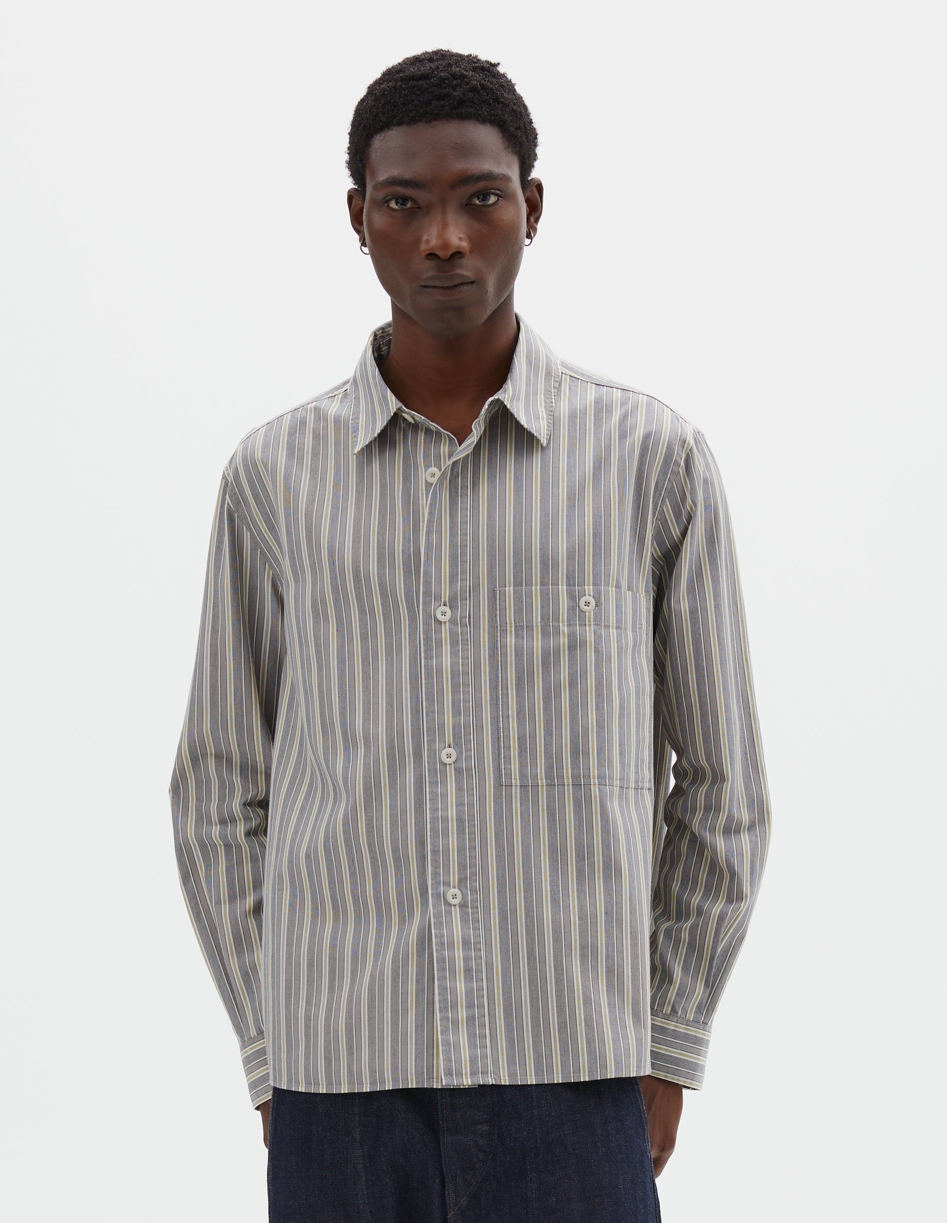 Cropped body image, front view of model wearing grey, white and yellow striped long sleeve cotton shirt, with dropped shoulder, raised placket, corozo brace button closure and button through chest patch pocket. Styled with indigo blue jeans.