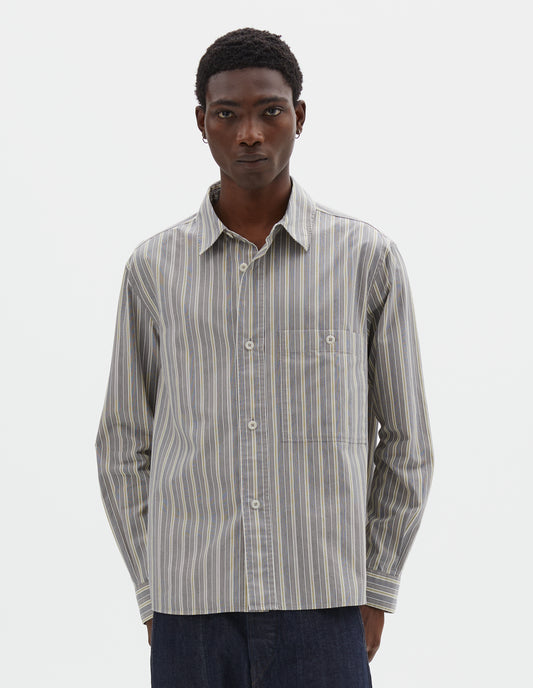 Cropped body image, front view of model wearing grey, white and yellow striped long sleeve cotton shirt, with dropped shoulder, raised placket, corozo brace button closure and button through chest patch pocket. Styled with indigo blue jeans.