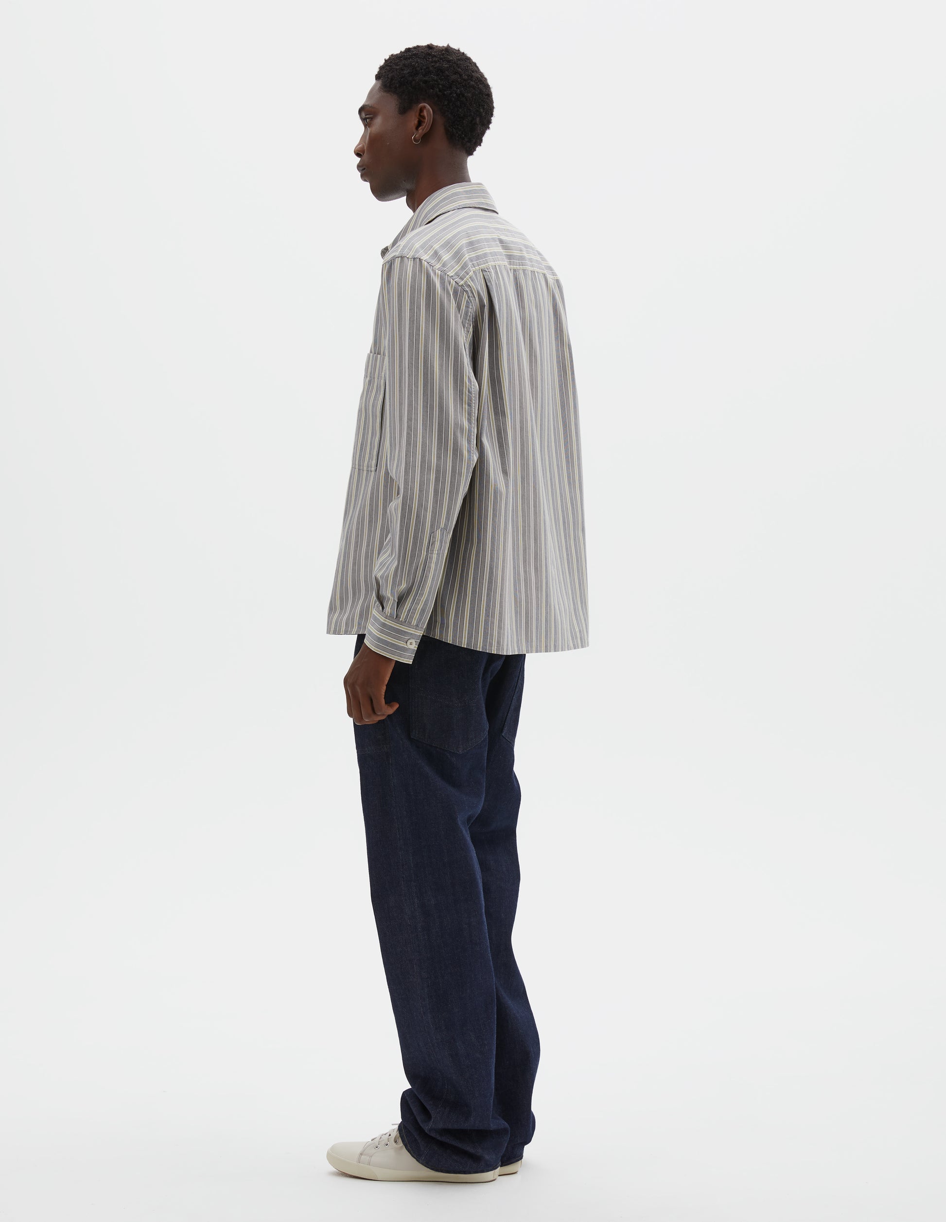 Full body image, back view of model wearing grey, white and yellow striped long sleeve cotton shirt. With dropped shoulder, corozo brace button cuffs and pleats on back yoke. Styled with indigo blue jeans and beige canvas trainers.