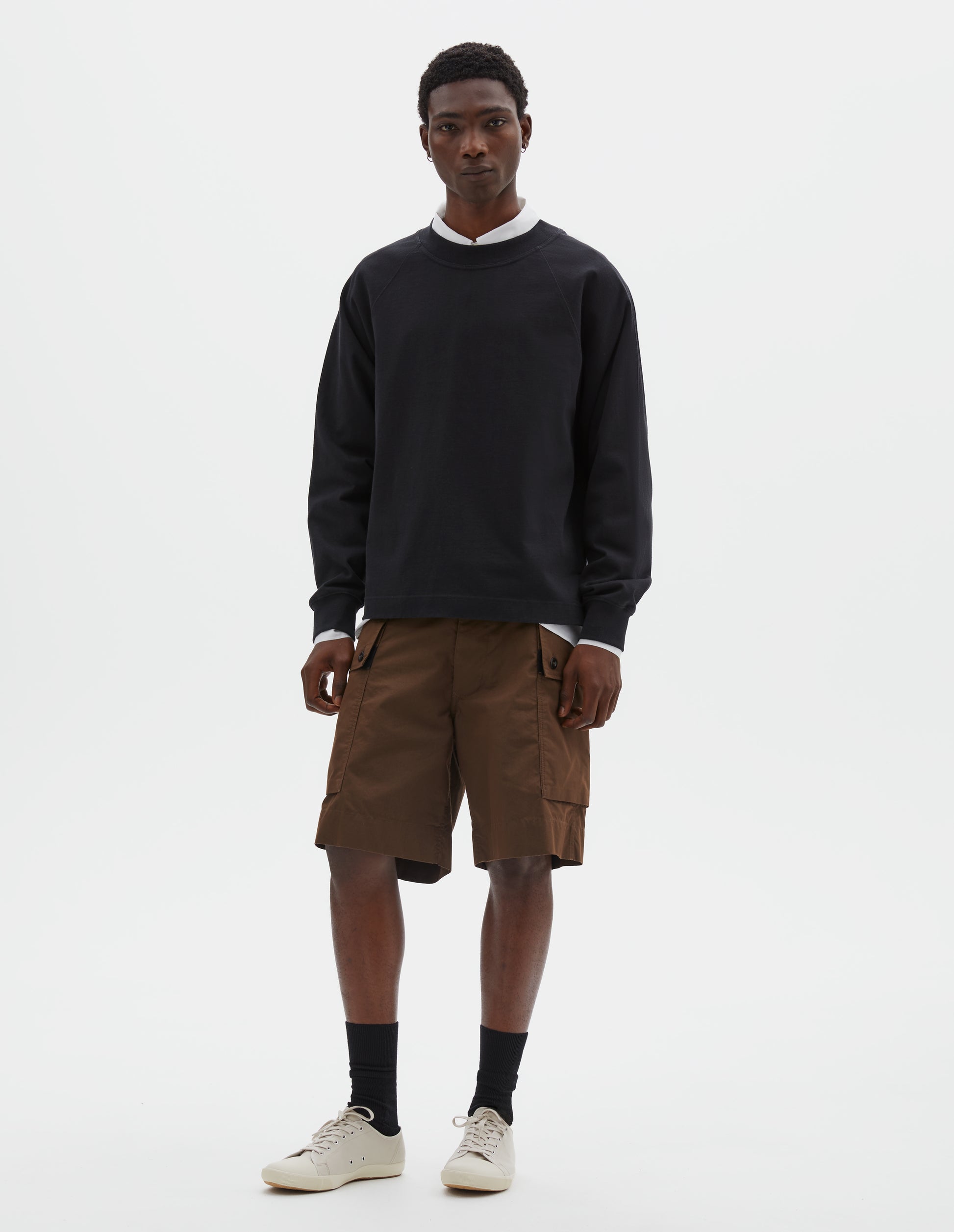 Full body image, front view of model wearing black cotton long sleeve t-shirt with raglan sleeve, single rib trims on neck and cuffs and vent split hem. Styled over white cotton shirt, brown shorts, with black socks and beige canvas trainers.