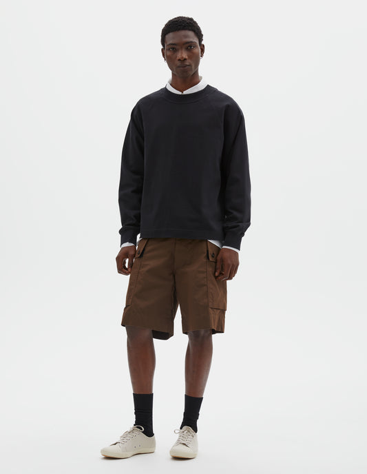 Full body image, front view of model wearing black cotton long sleeve t-shirt with raglan sleeve, single rib trims on neck and cuffs and vent split hem. Styled over white cotton shirt, brown shorts, with black socks and beige canvas trainers.