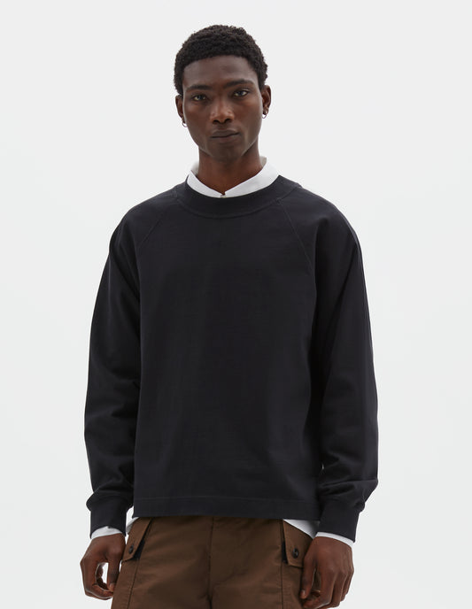 Cropped body image, front view of model wearing black cotton long sleeve t-shirt with raglan sleeve, single rib trims on neck and cuffs and vent split hem. Styled over white cotton shirt with brown shorts.