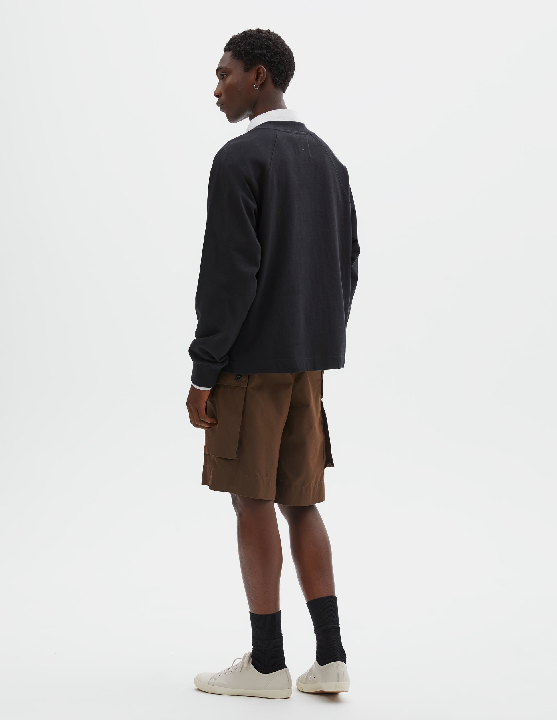 Full body image, back view of model wearing black cotton long sleeve t-shirt with raglan sleeve, single rib trims on neck and cuffs and vent split hem. Styled over white cotton shirt, brown shorts, with black socks and beige canvas trainers.