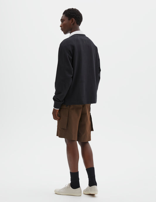 Full body image, back view of model wearing black cotton long sleeve t-shirt with raglan sleeve, single rib trims on neck and cuffs and vent split hem. Styled over white cotton shirt, brown shorts, with black socks and beige canvas trainers.