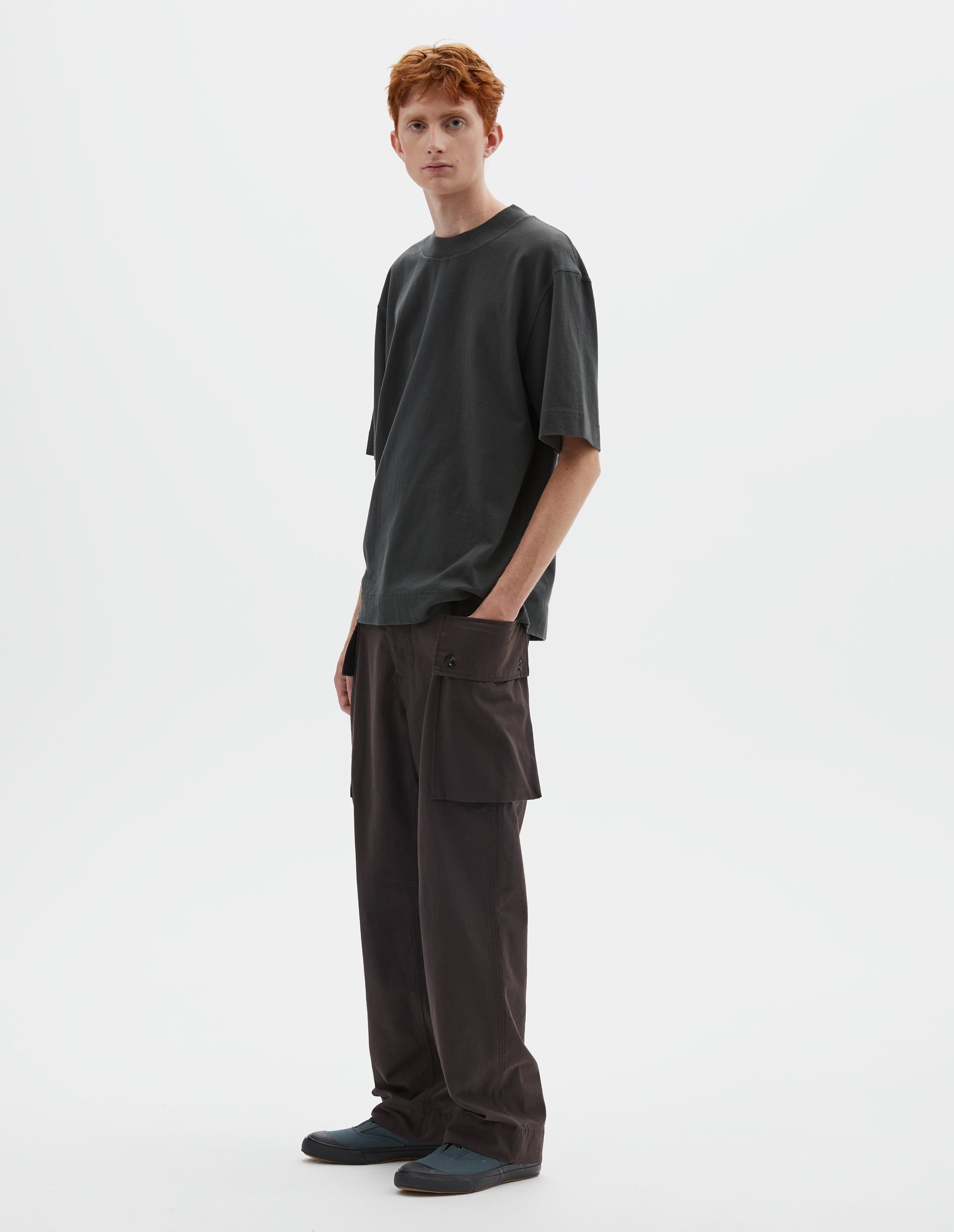 Full body image, front view of model wearing greenish black short sleeve t-shirt with dropped shoulder and rib neck. Styled with reddish brown cargo trousers and blue canvas trainers.
