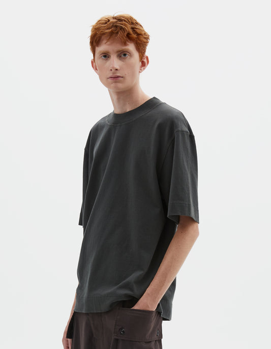 Cropped body image, front view of model wearing greenish black short sleeve t-shirt with dropped shoulder and rib neck. Styled with reddish brown cargo trousers.