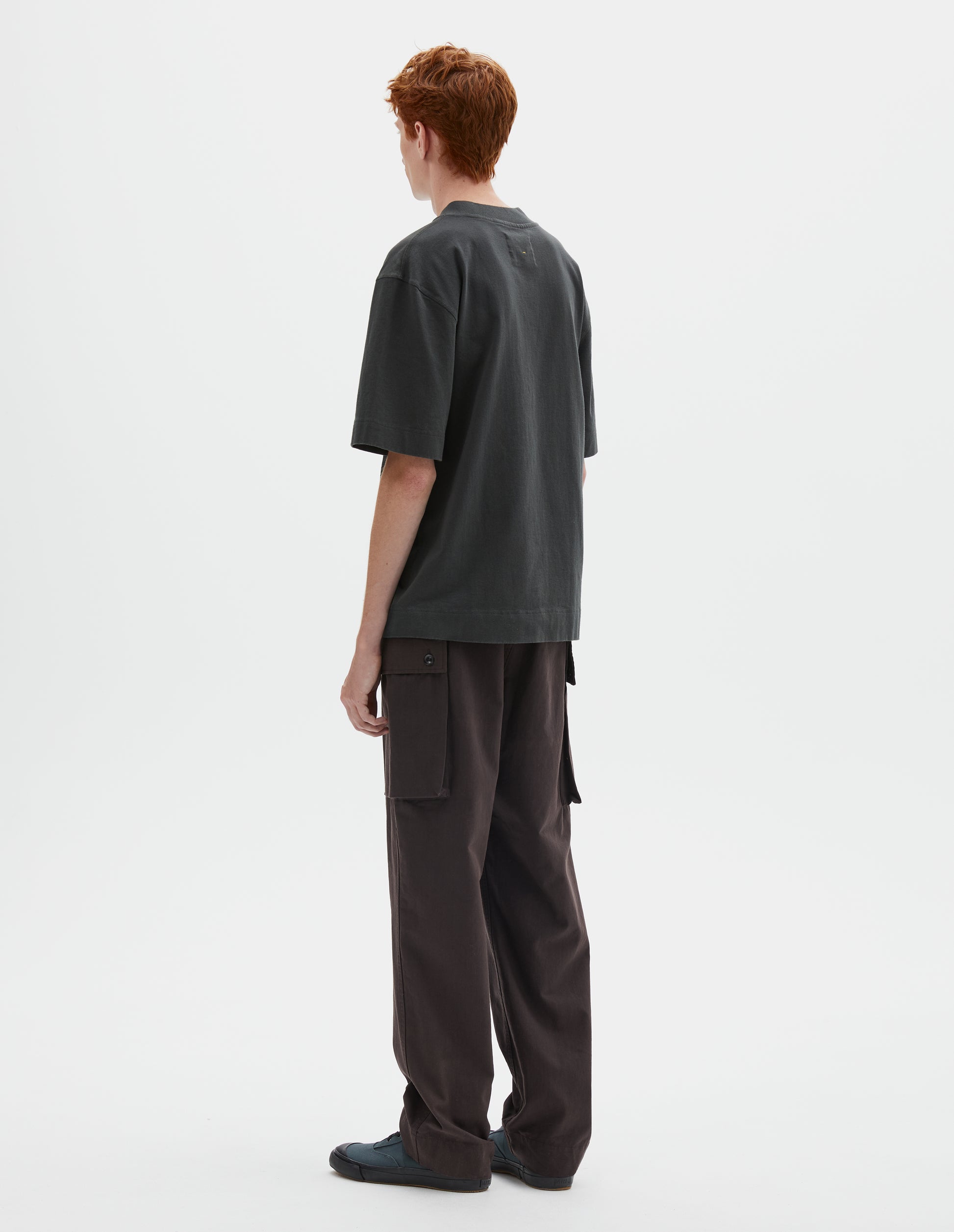 Full body image, back view of model wearing greenish black short sleeve t-shirt with dropped shoulder and rib neck. Styled with reddish brown cargo trousers and blue canvas trainers.
