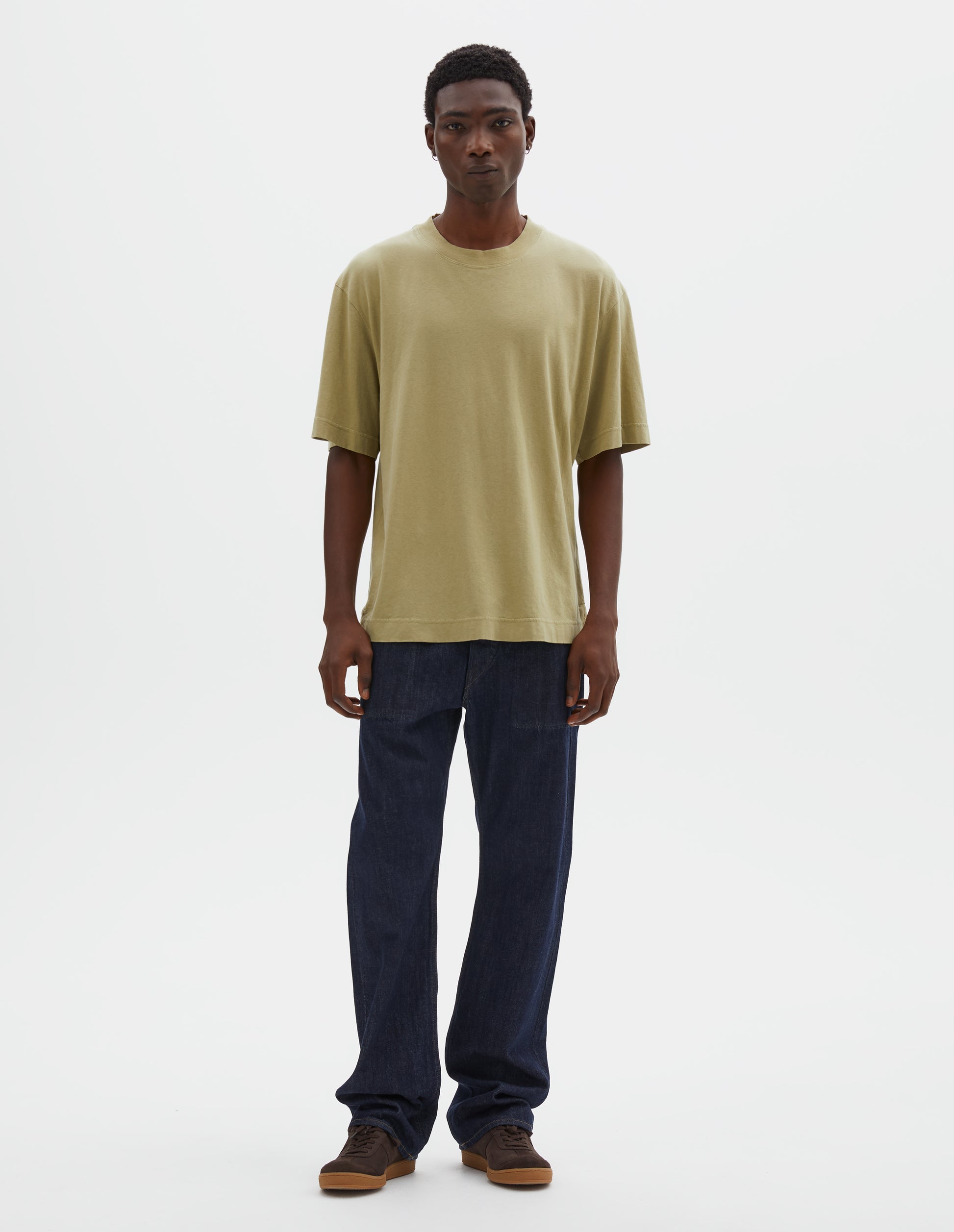 Full body image, front view of model wearing pale green cotton linen short sleeve t-shirt with vent split hem. Styled with indigo blue jeans and brown suede trainers.