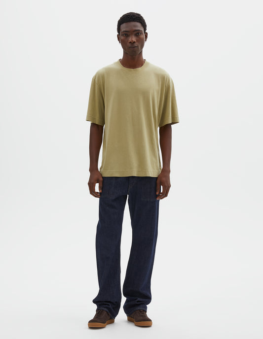 Full body image, front view of model wearing pale green cotton linen short sleeve t-shirt with vent split hem. Styled with indigo blue jeans and brown suede trainers.