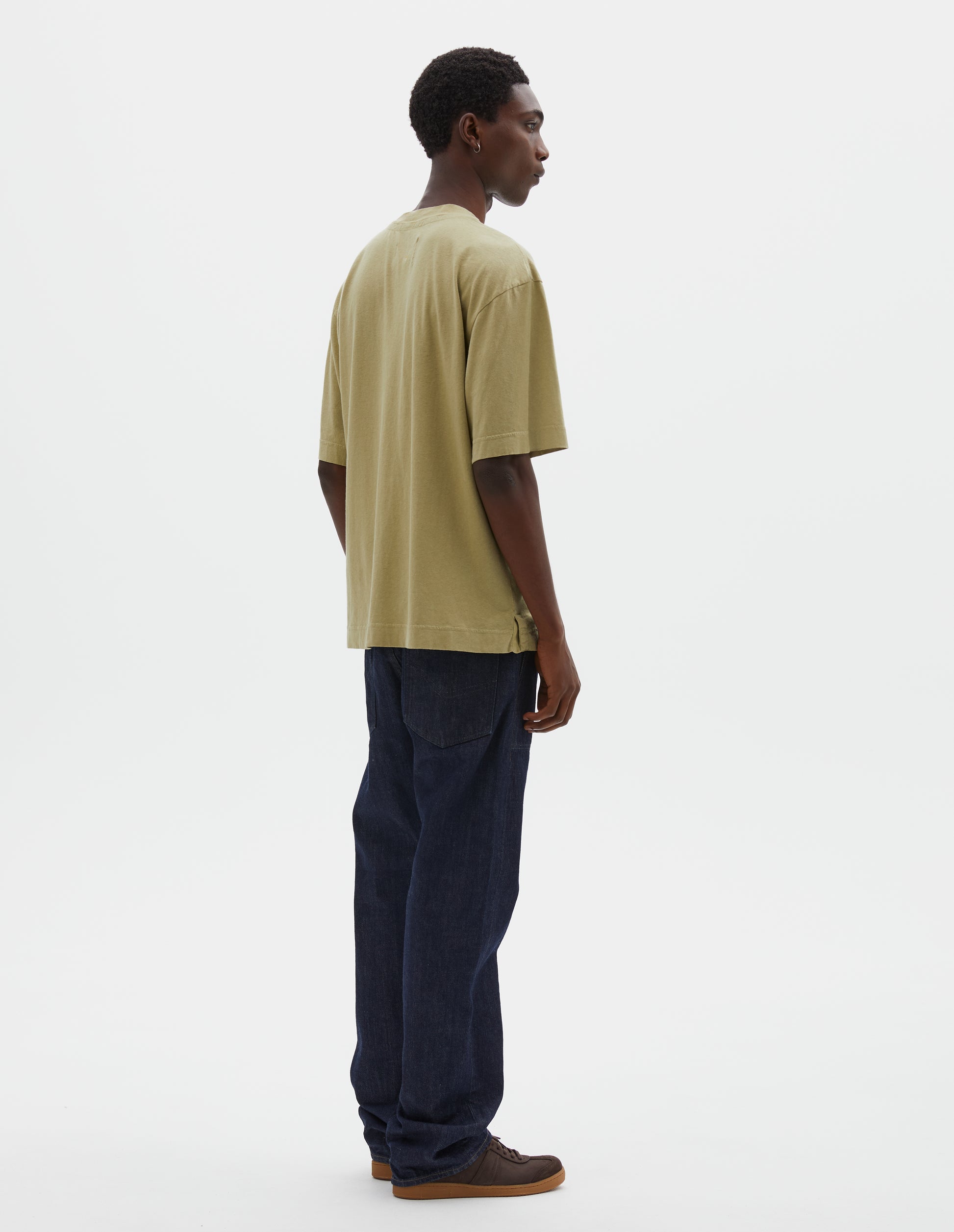 Full body image, back view of model wearing pale green cotton linen short sleeve t-shirt with vent split hem. Styled with indigo blue jeans and brown suede trainers.