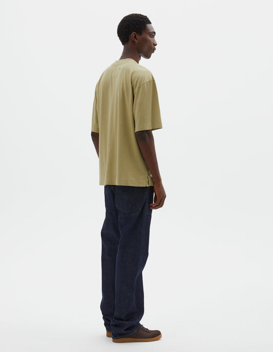Full body image, back view of model wearing pale green cotton linen short sleeve t-shirt with vent split hem. Styled with indigo blue jeans and brown suede trainers.
