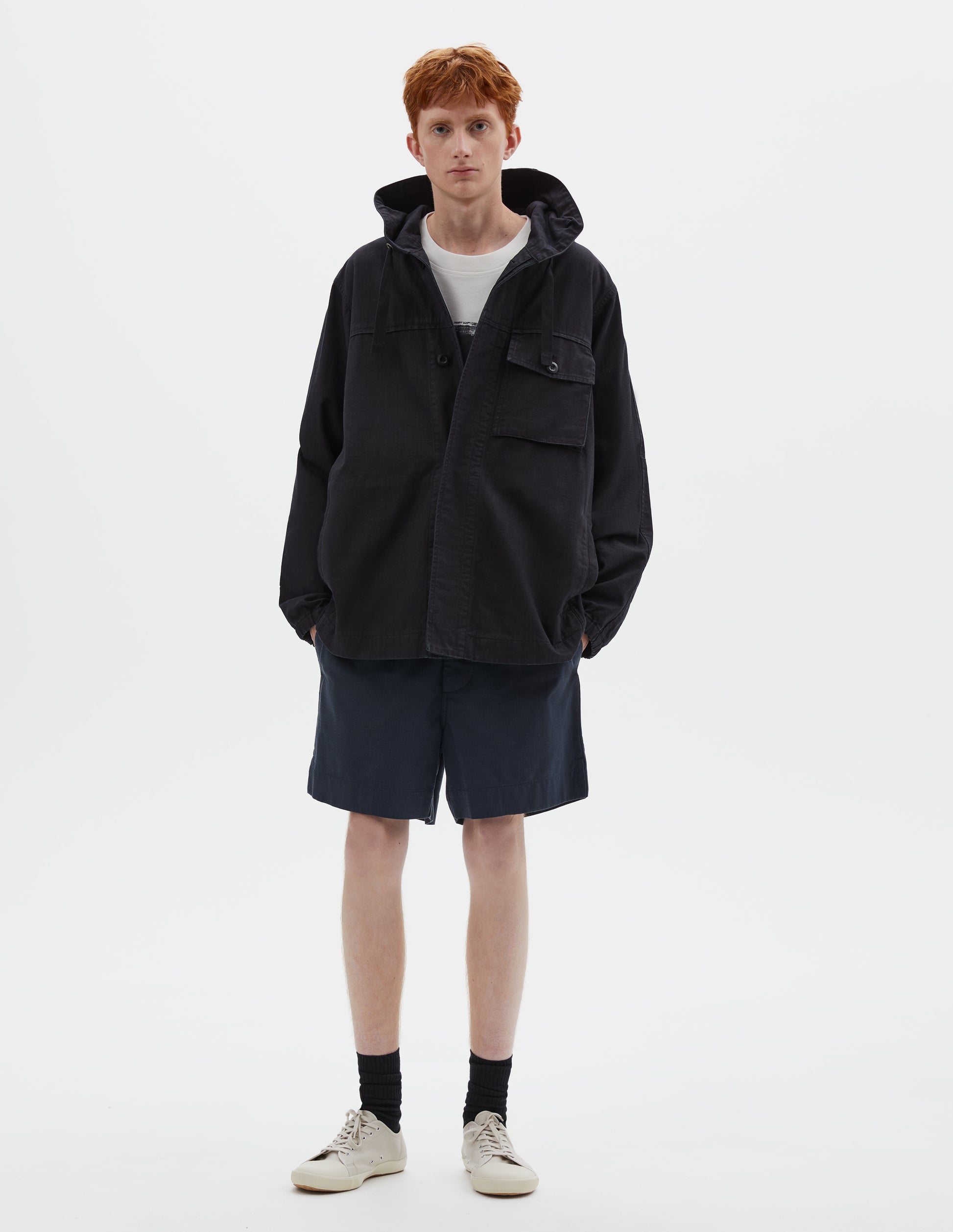 Full body image, front view of model wearing indigo blue twill hooded jacket, with flap patch chest pocket, concealed placket, drawcord adjustable hood and elasticated cuffs. Styled over white graphic t-shirt, with navy blue shorts, black socks and beige canvas trainers.