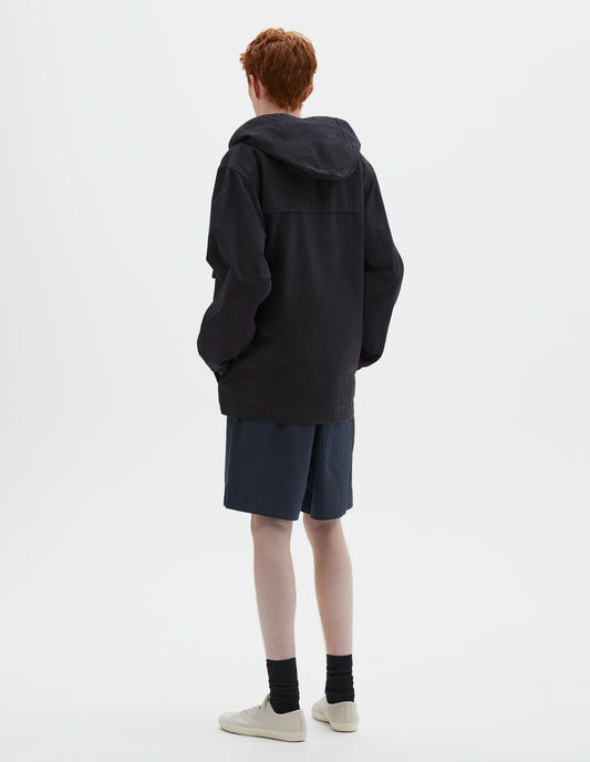 Full body image, back view of model wearing indigo blue twill hooded jacket, with sleeve patches. Styled with navy blue shorts, black socks and beige canvas trainers.
