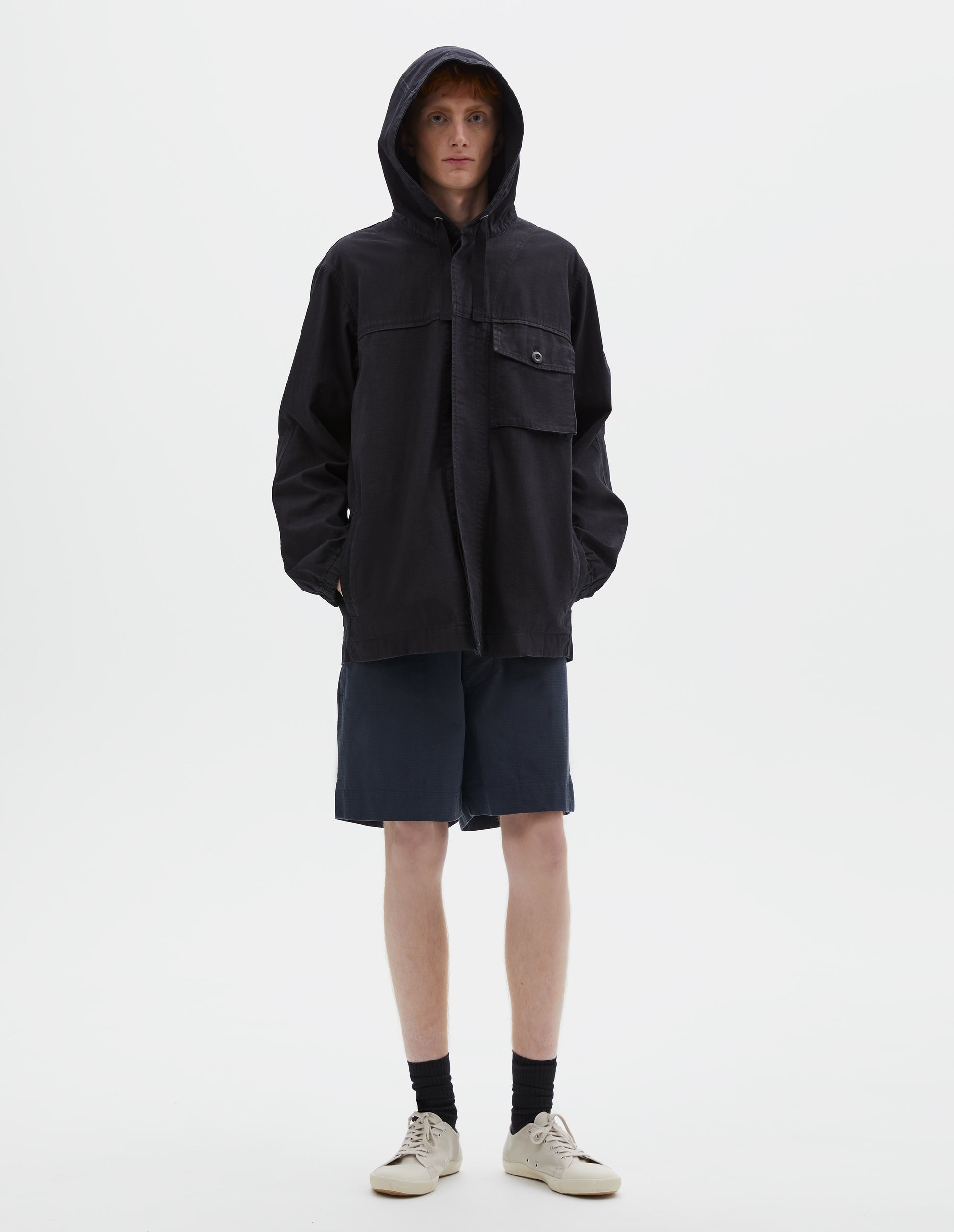 Full body image, front view of model wearing indigo blue twill hooded jacket, with flap patch chest pocket, concealed placket, drawcord adjustable hood styled over head. Styled with navy blue shorts, black socks and beige canvas trainers.