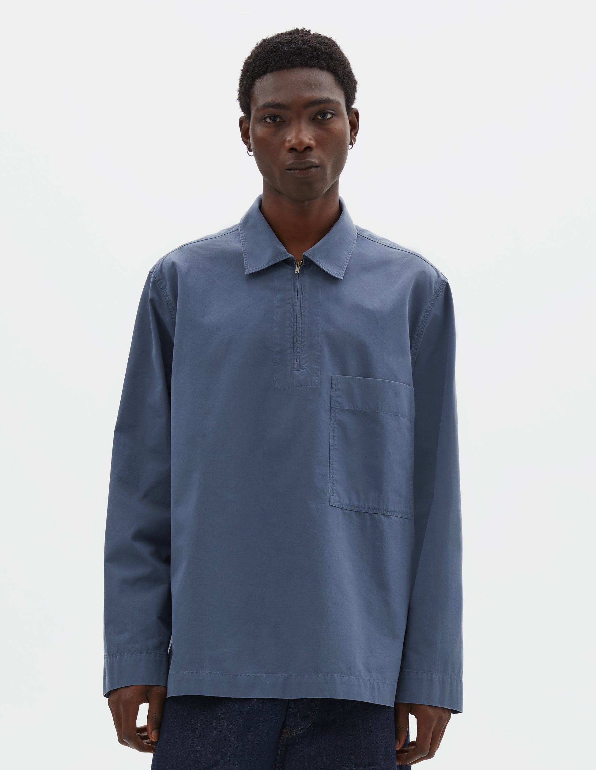 Cropped body image, front view of model wearing relaxed blue shirt with wide stitch-through placket with half zip, large patch chest pocket, straight cuffs and and side split hem. Styled with dark blue jean shorts.