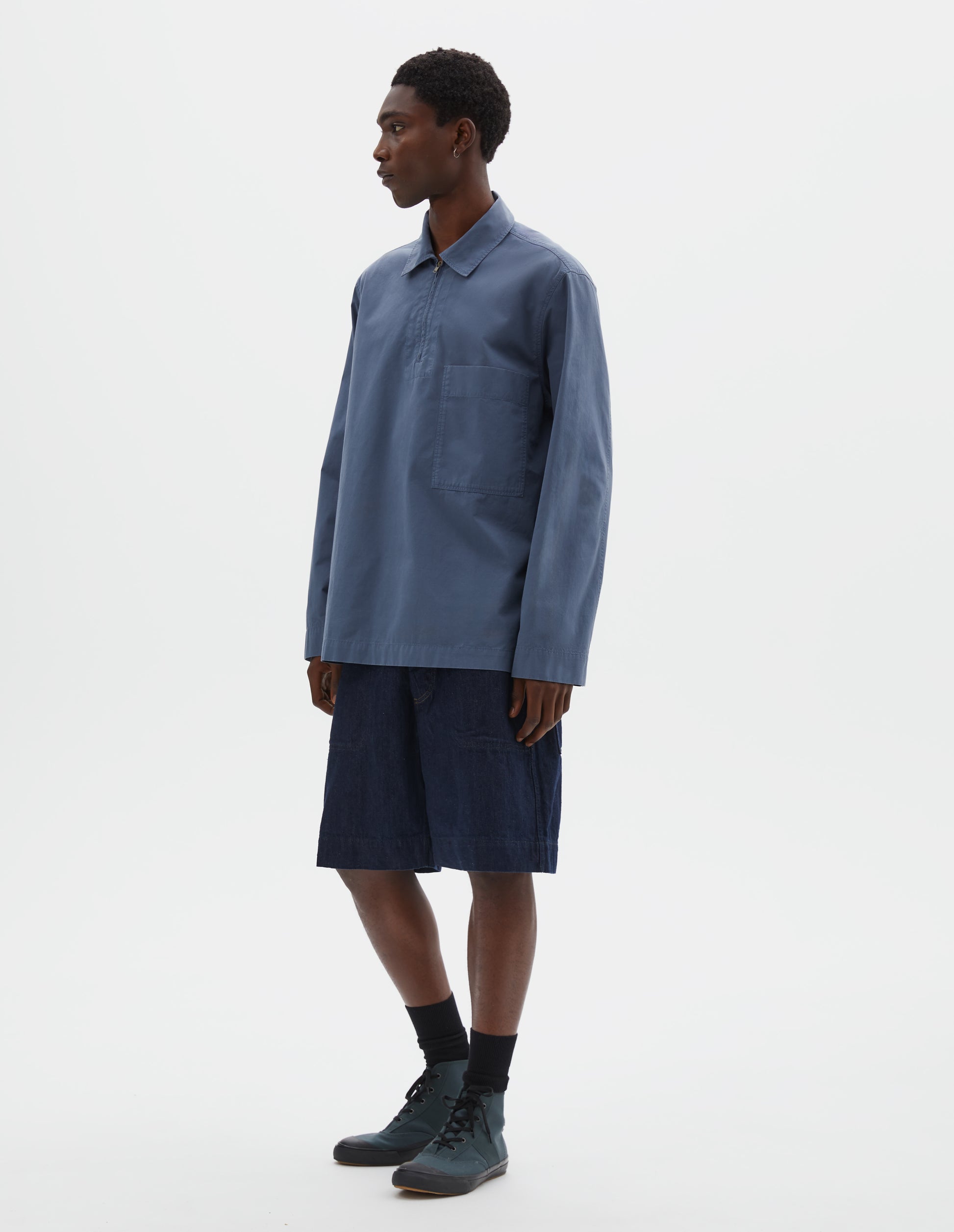 Full body image, front view of model wearing relaxed blue shirt with wide stitch-through placket with half zip, large patch chest pocket, straight cuffs and and side split hem. Styled with dark blue jean shorts, black socks and green high top trainers.
