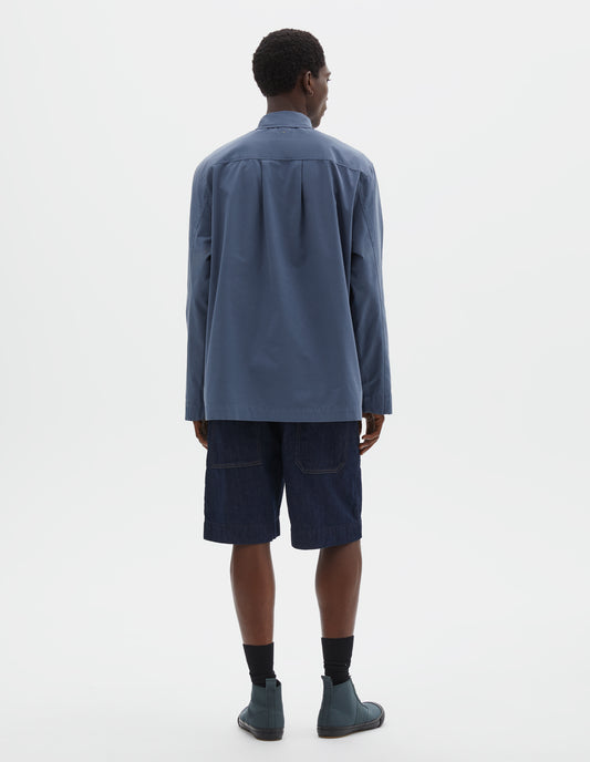Full body image, back view of model wearing relaxed blue shirt with straight cuffs, side split hem and box pleat on back yoke. Styled with dark blue jean shorts, black socks and green high top trainers.