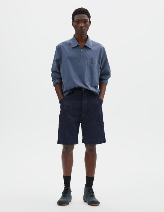 Full body image, front view of model wearing indigo blue denim twill shorts. With metal button tab waist adjusters, metal button fly, belt loops and slant stitch-through pockets. Styled with blue shirt, black socks and blue canvas trainers.