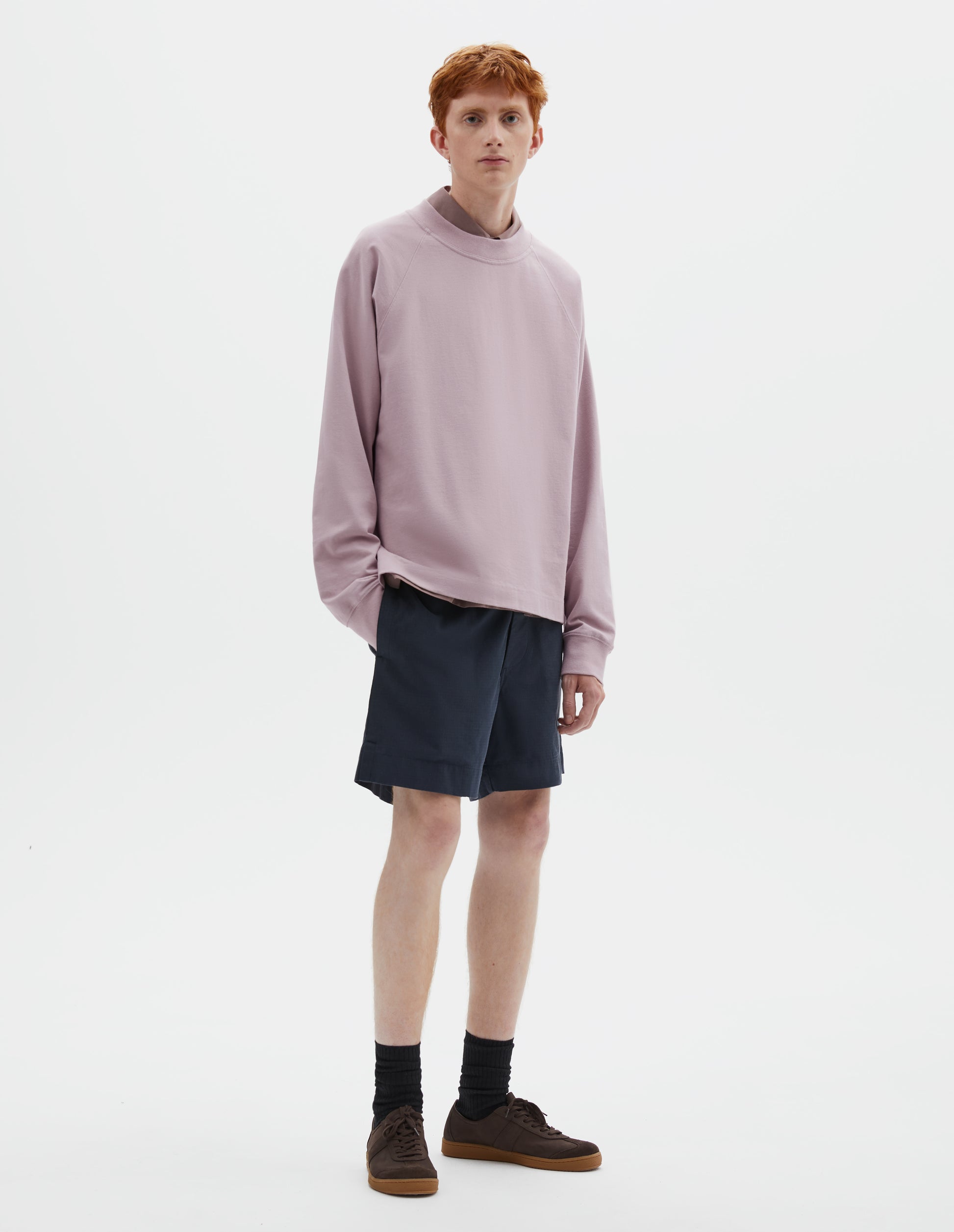 Full body image, front view of model wearing pink cotton long sleeve t-shirt with raglan sleeve and single rib trims on neck and cuffs. Styled over dark pink shirt, dark blue shorts and black socks with dark brown nubuck trainers.
