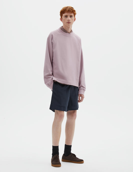 Full body image, front view of model wearing pink cotton long sleeve t-shirt with raglan sleeve and single rib trims on neck and cuffs. Styled over dark pink shirt, dark blue shorts and black socks with dark brown nubuck trainers.