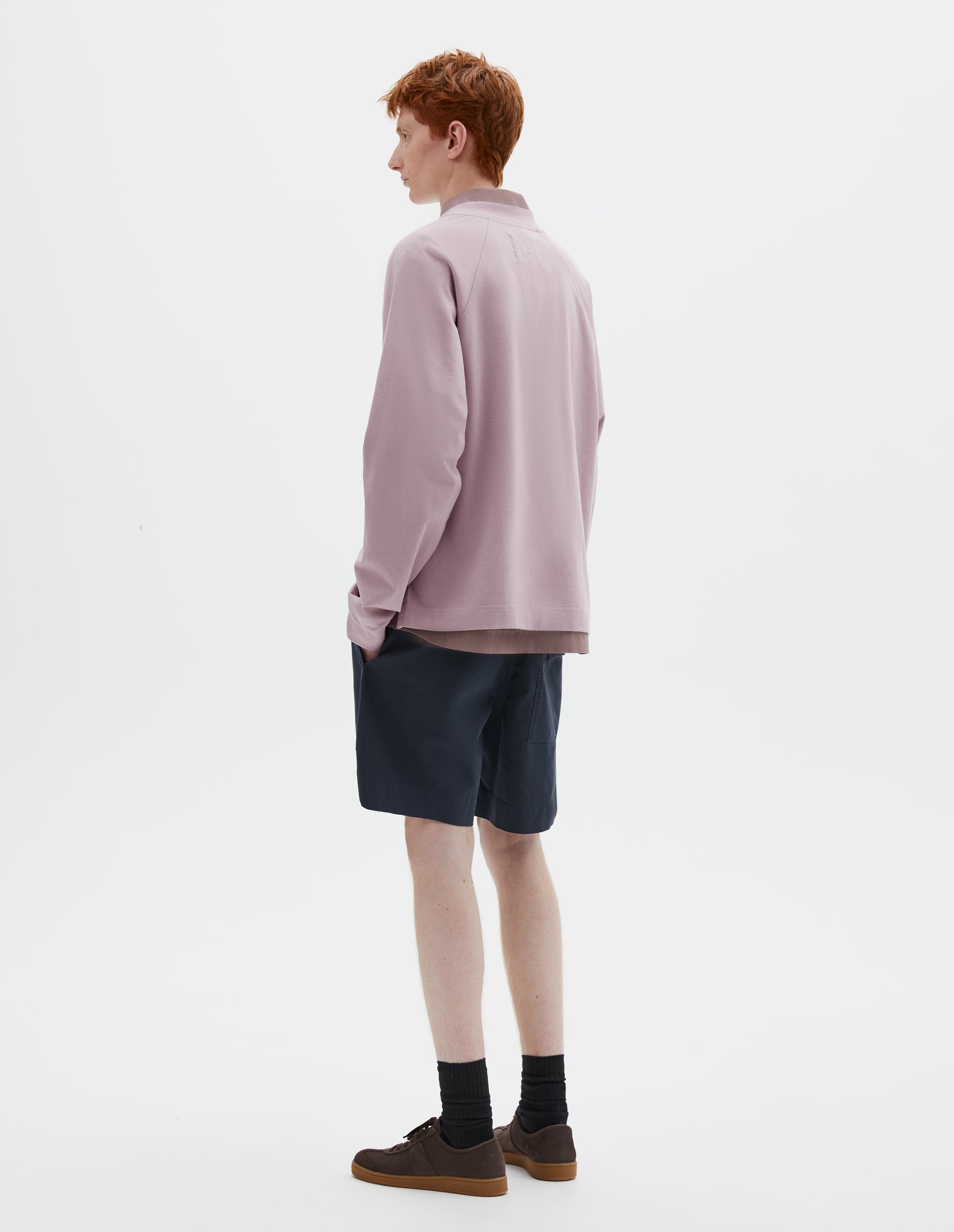 Full body image, back view of model wearing pink cotton long sleeve t-shirt with raglan sleeve, single rib trims on neck and cuffs, and vent split hem. Styled over dark pink shirt, dark blue shorts and black socks with dark brown nubuck trainers.