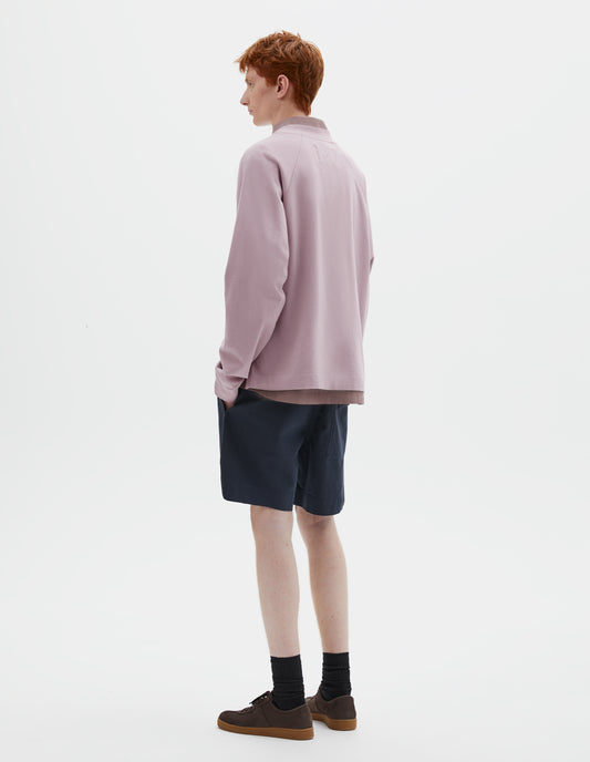 Full body image, back view of model wearing pink cotton long sleeve t-shirt with raglan sleeve, single rib trims on neck and cuffs, and vent split hem. Styled over dark pink shirt, dark blue shorts and black socks with dark brown nubuck trainers.