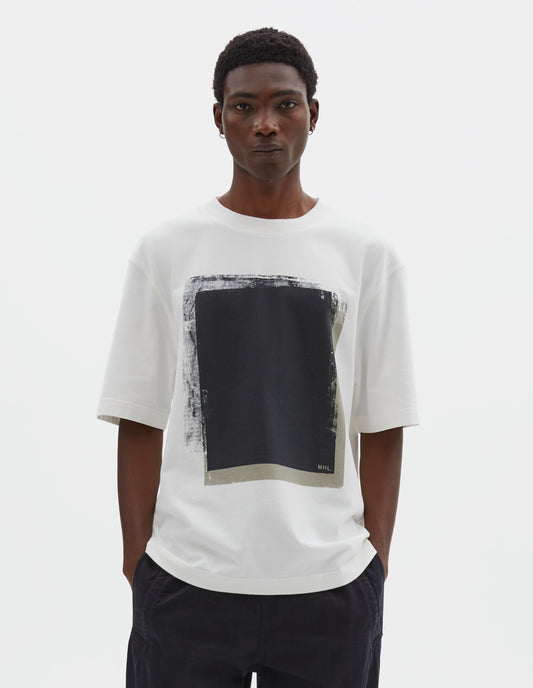 Cropped body image, front view of model wearing off white short sleeve t shirt with black abstract block printed on the front. Styled with black trousers.