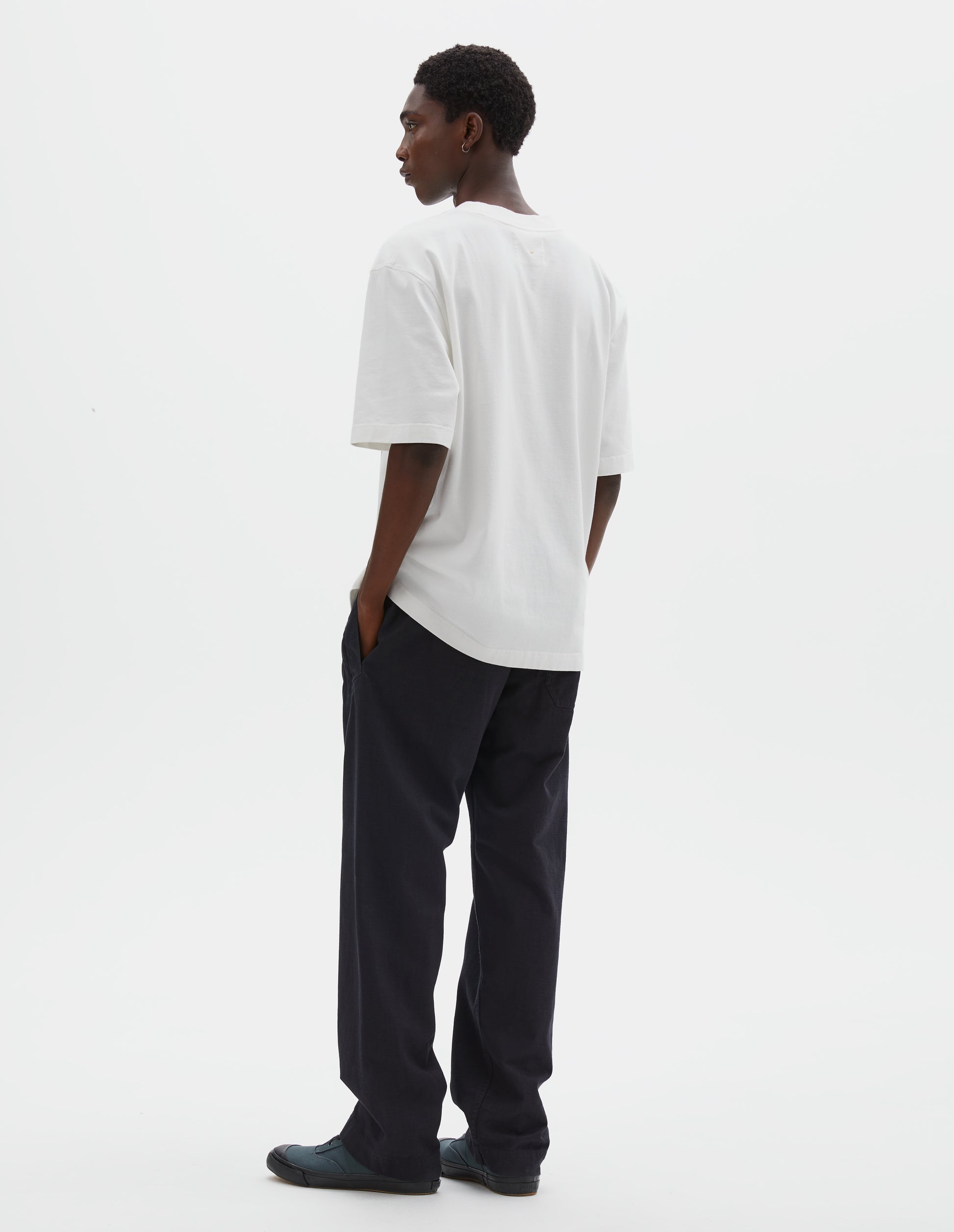 Full body image, back view of model wearing off white short sleeve t shirt. Styled with black trousers and blue canvas trainers.