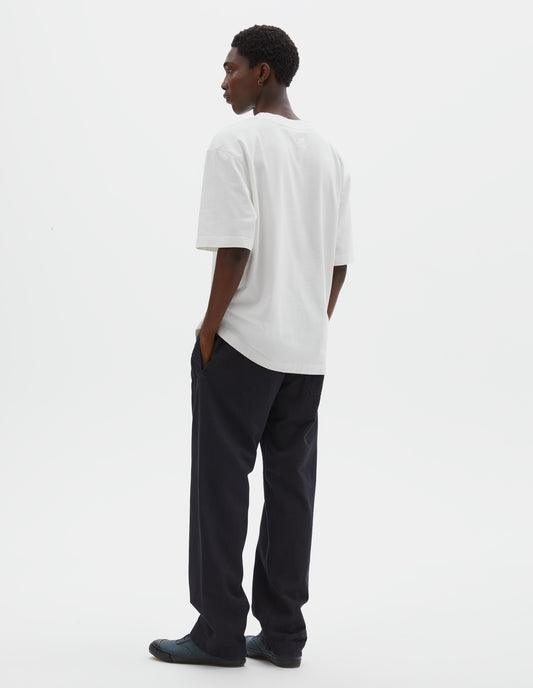 Full body image, back view of model wearing off white short sleeve t shirt. Styled with black trousers and blue canvas trainers.